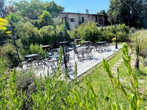 Un'area salotto all'aperto con tavoli e sedie, immersa nel verde e con un edificio in pietra sullo sfondo.