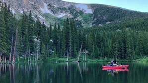 Un lago tranquilo con un kayak rojo y un velero, rodeado de un bosque denso y montañas.
