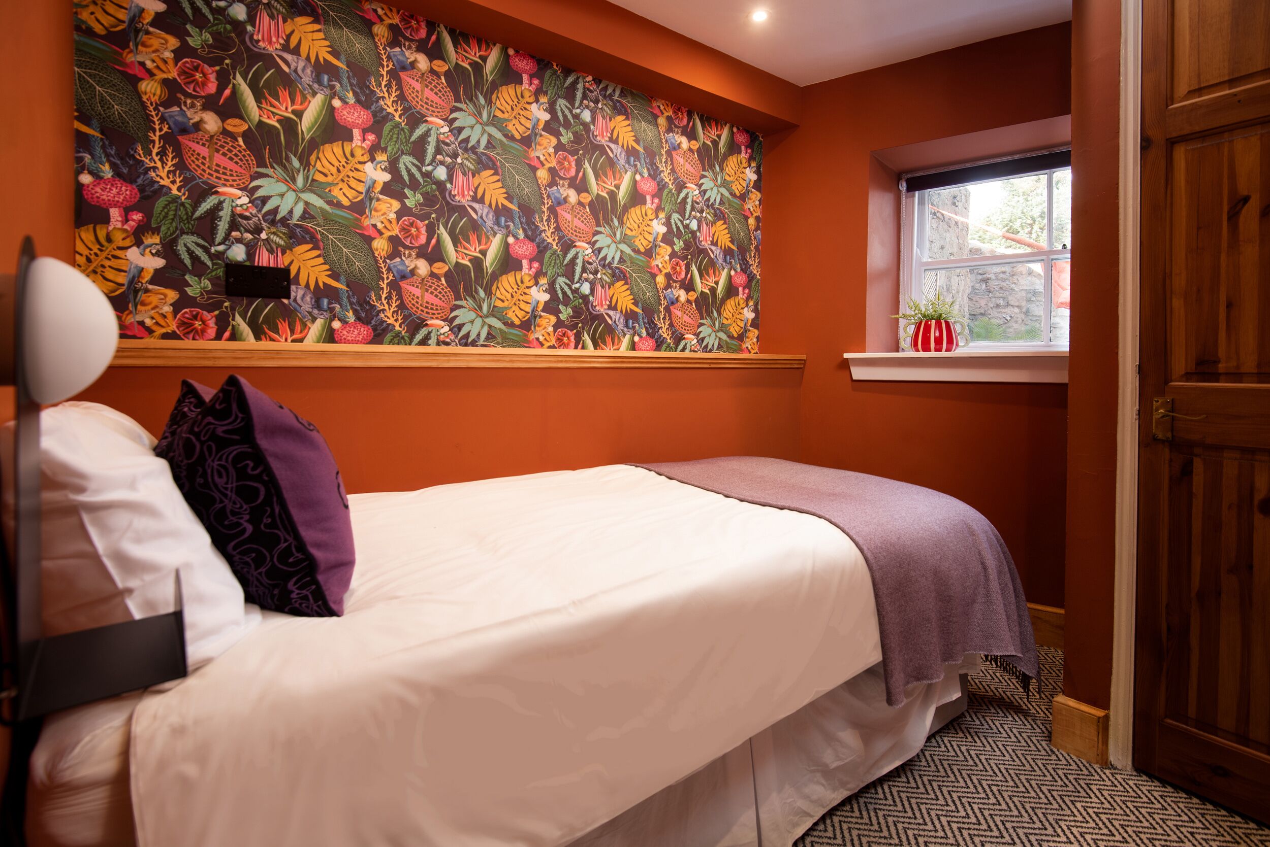 Une chambre avec un lit, une fenêtre donnant sur un beau paysage et un mur décoré d’une fresque florale aux couleurs vives.
