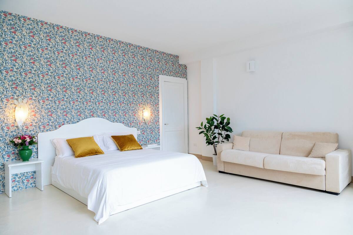 Un dormitorio con cama, mesita de noche, un florero verde con flores, sofá color beige y una pared con papel tapiz floral.