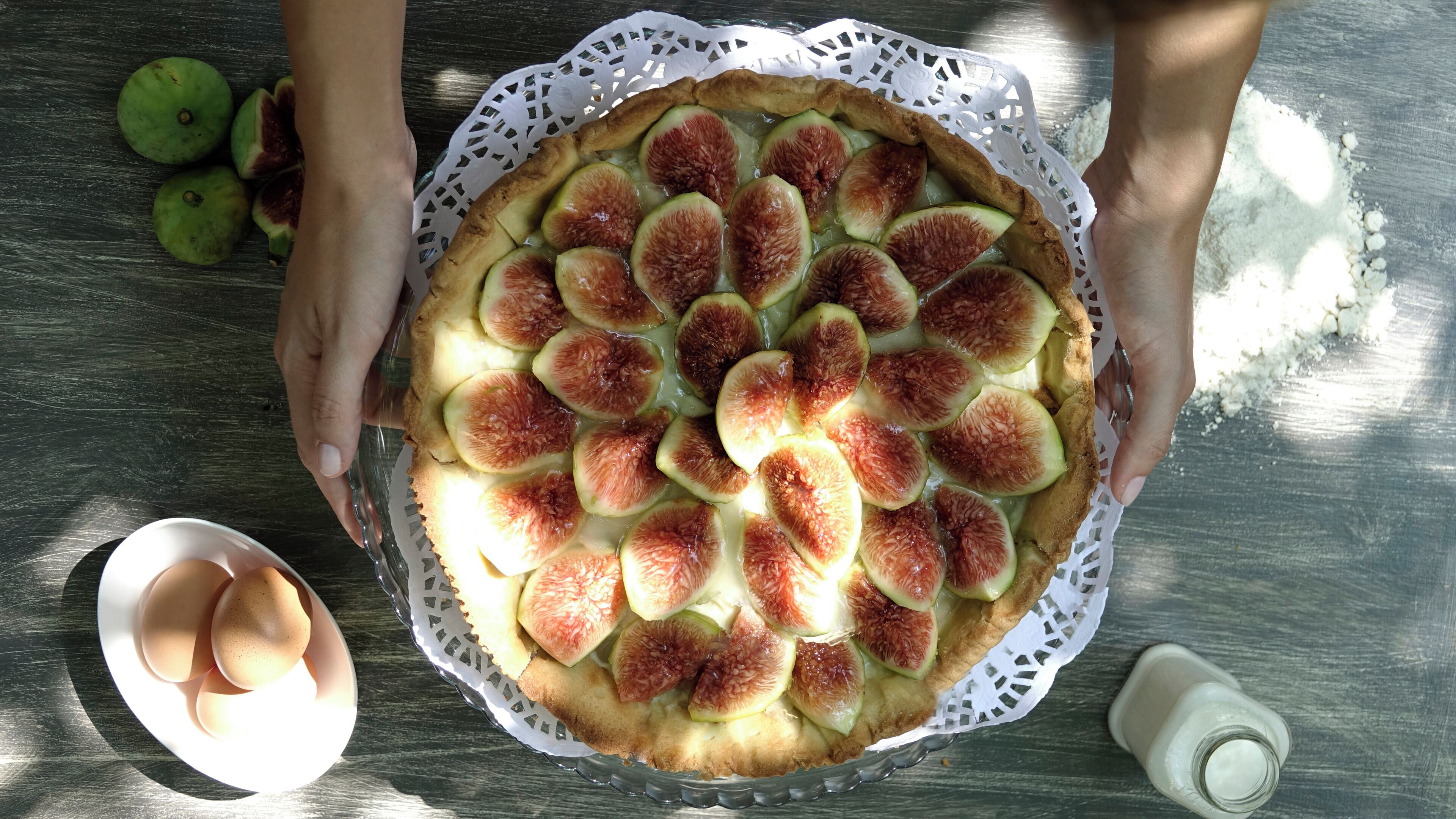 Une tarte garnie de tranches de figues, posée sur une nappe blanche en dentelle, avec des œufs et une bouteille sur une table en bois.