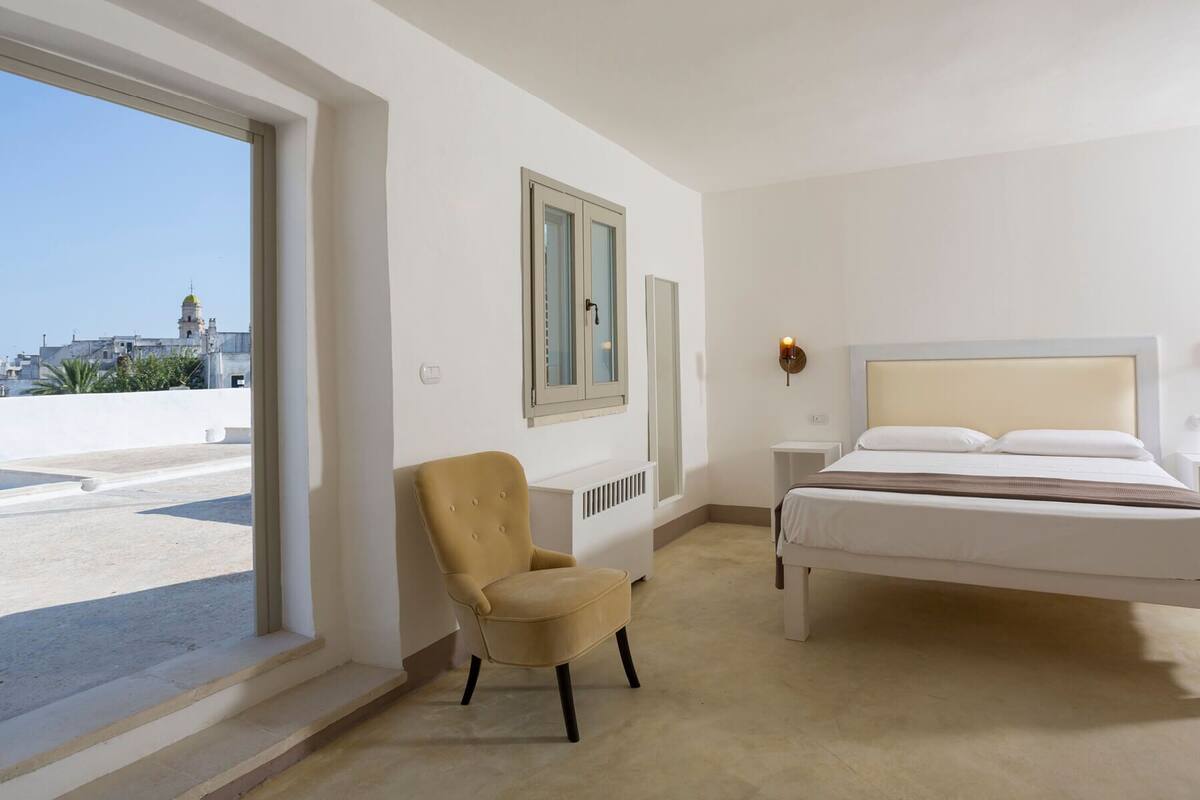 Un dormitorio con cama, una silla, un ventanal y vistas a un edificio.