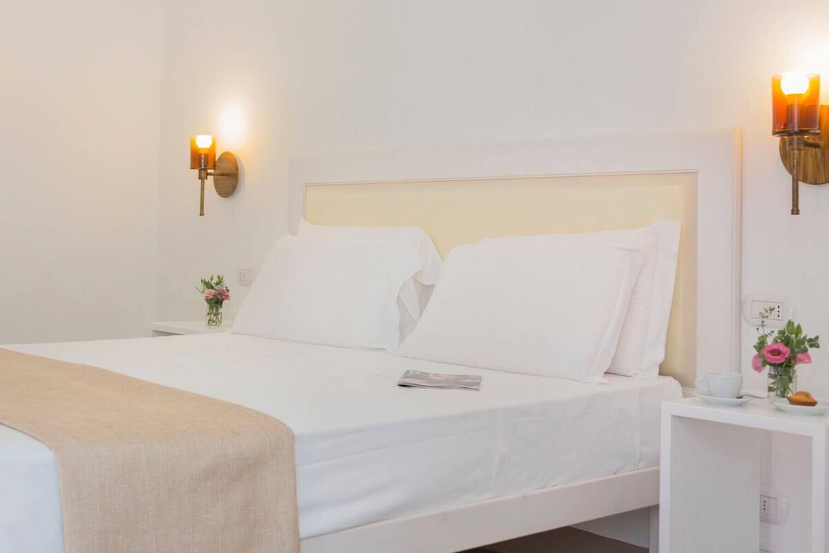 Una cama bien tendida con sábanas blancas, una mesita de noche con una taza y un florero, y lámparas fijadas a la pared.