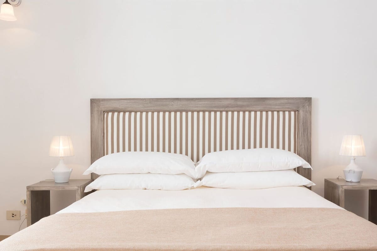 Una cama con cabecera de madera, dos mesitas de noche con lámparas y una pared lisa.