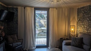 Un salon comprenant un canapé, un fauteuil, une table basse, un téléviseur fixé au mur, une fenêtre avec des rideaux, un escalier extérieur et un luminaire décoratif.