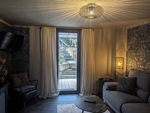 Un soggiorno con divano, una poltrona, tavolino, televisione a muro, finestra con tende e una scala esterna, oltre a un lampadario decorativo.