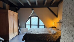 Une chambre avec un plafond en pente, une grande fenêtre, une armoire en bois et une lampe de chevet.