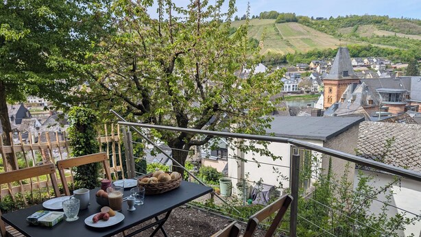 Un balcon avec une table dressée pour un repas, donnant sur un village aux bâtiments traditionnels et sur une colline en arrière-plan.