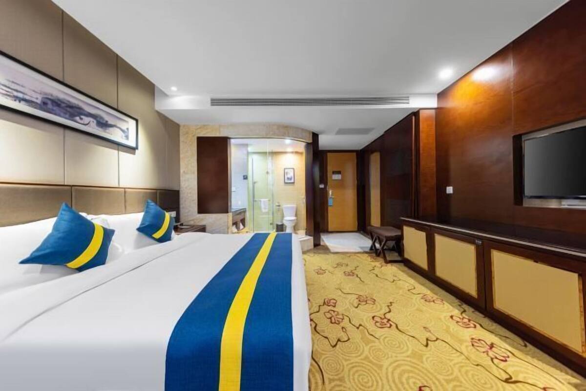 A hotel room with a bed, a television, a desk, and a bathroom visible through a glass partition.