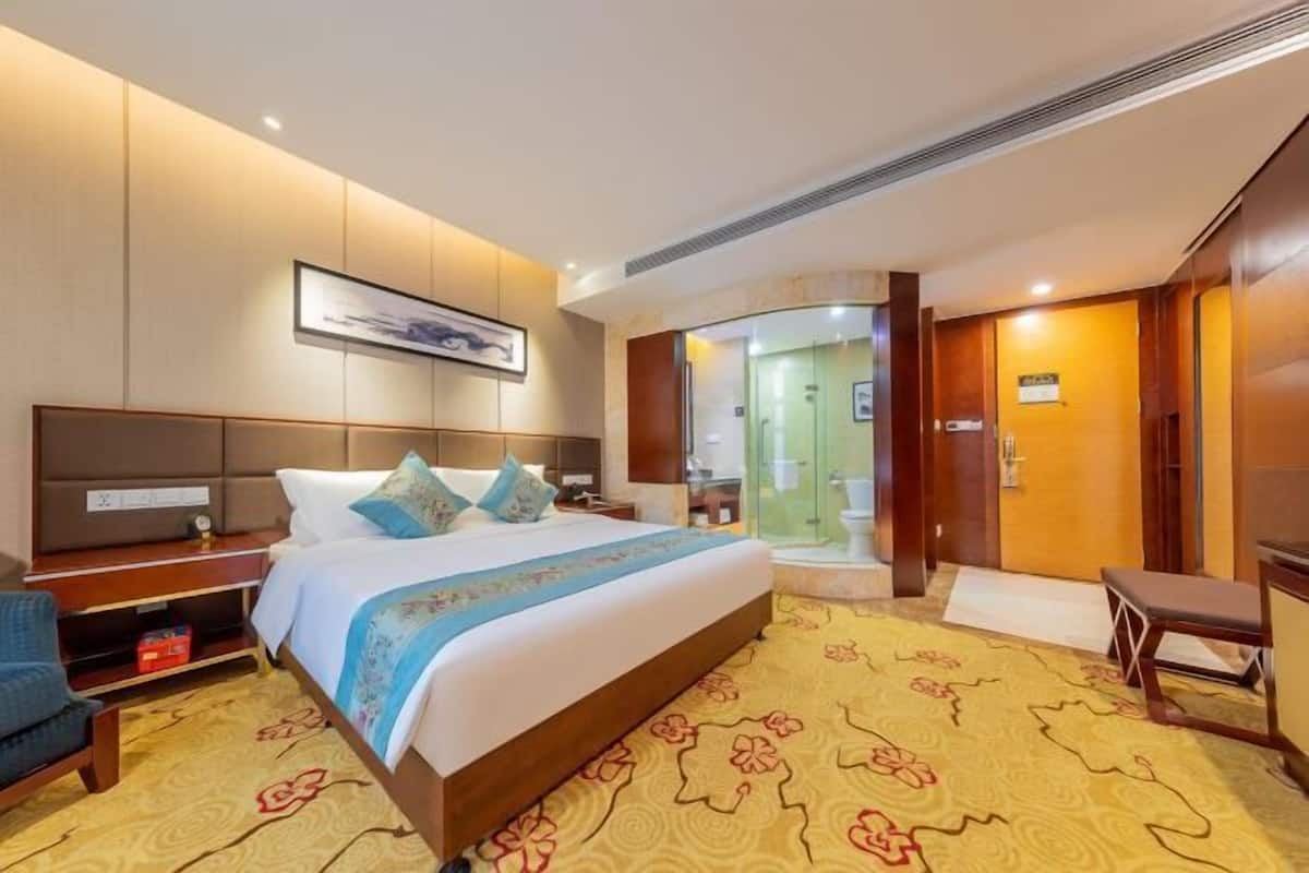 A hotel room with a large bed, a desk, a chair, and a bathroom visible through a glass door.