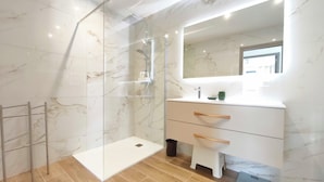 A modern bathroom with a glass shower enclosure, a marble-tiled floor, a white vanity with a mirror, and a wooden towel rack.