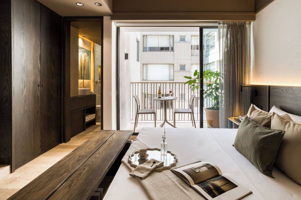 A hotel room with a bed, a bedside table, a small balcony with a table and chairs, and a view of the cityscape.