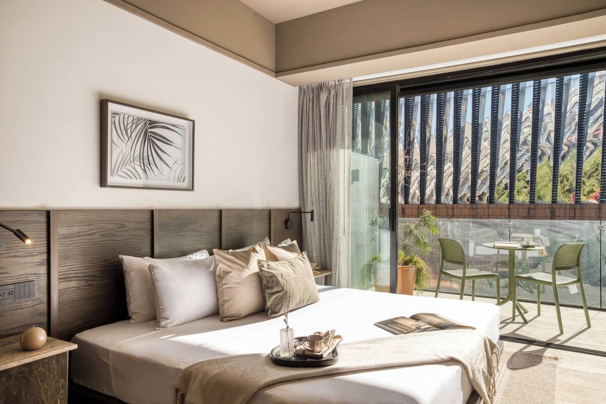 A modern hotel room with a large bed, a bedside table with a lamp, a framed artwork on the wall, and a balcony with chairs and a table.