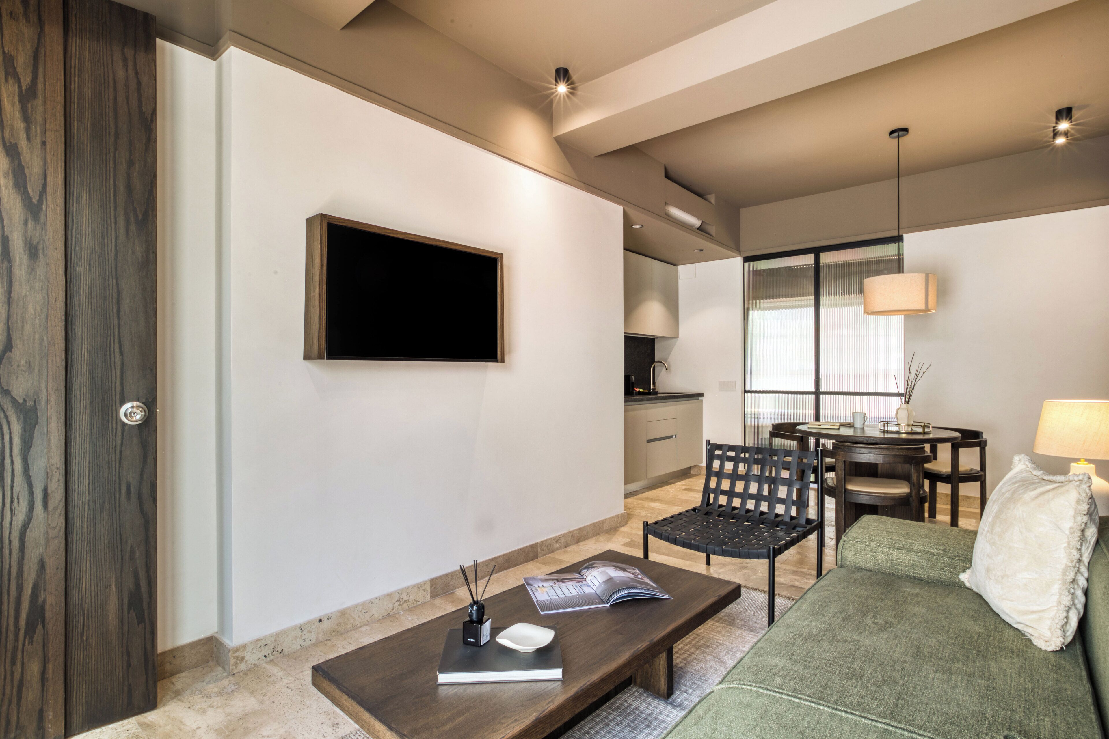 A modern living room with a flat-screen TV, a coffee table with a book and a remote, a green sofa, and a dining area with a lamp.