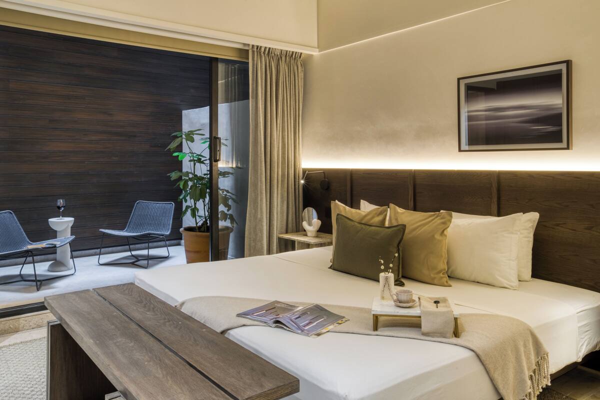 A hotel room with a large bed, a wooden bench, a small table with a cup and a book, a potted plant, and a view of the outdoors through a sliding door.