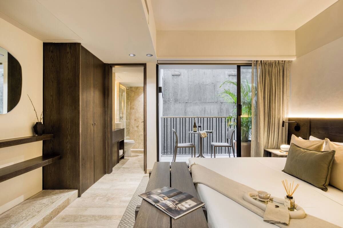 A modern hotel room with a large bed, a small balcony with a table and chairs, and a view of the city.
