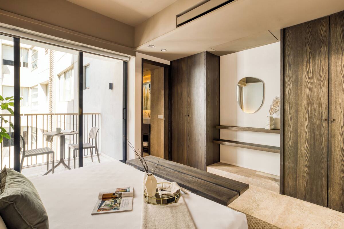 A modern bedroom with a large bed, a small table with a vase, and a view of the outside balcony.