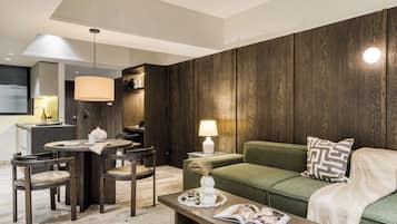 A modern living room with a green sofa, wooden panel walls, a dining area with a table and chairs, and a kitchen area in the background.