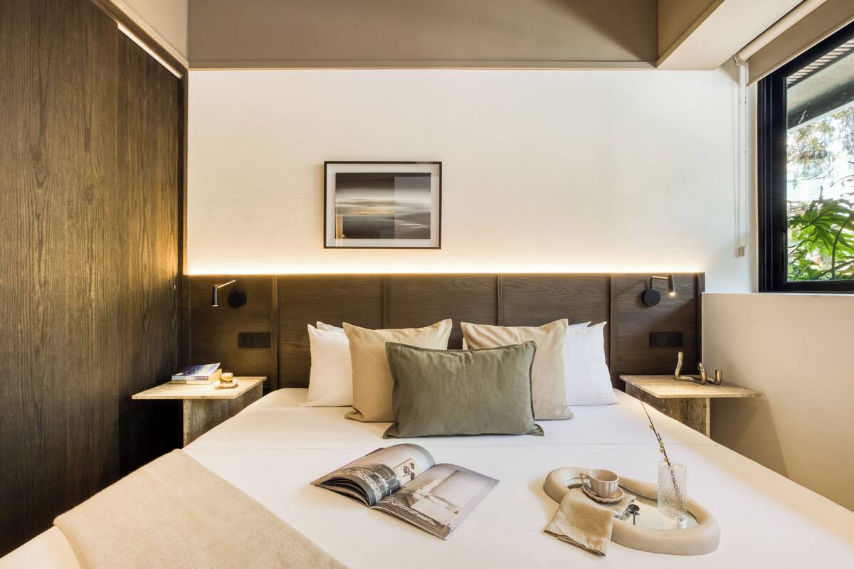 A modern hotel room with a large bed, a bedside table with a magazine, a cup, and a vase.