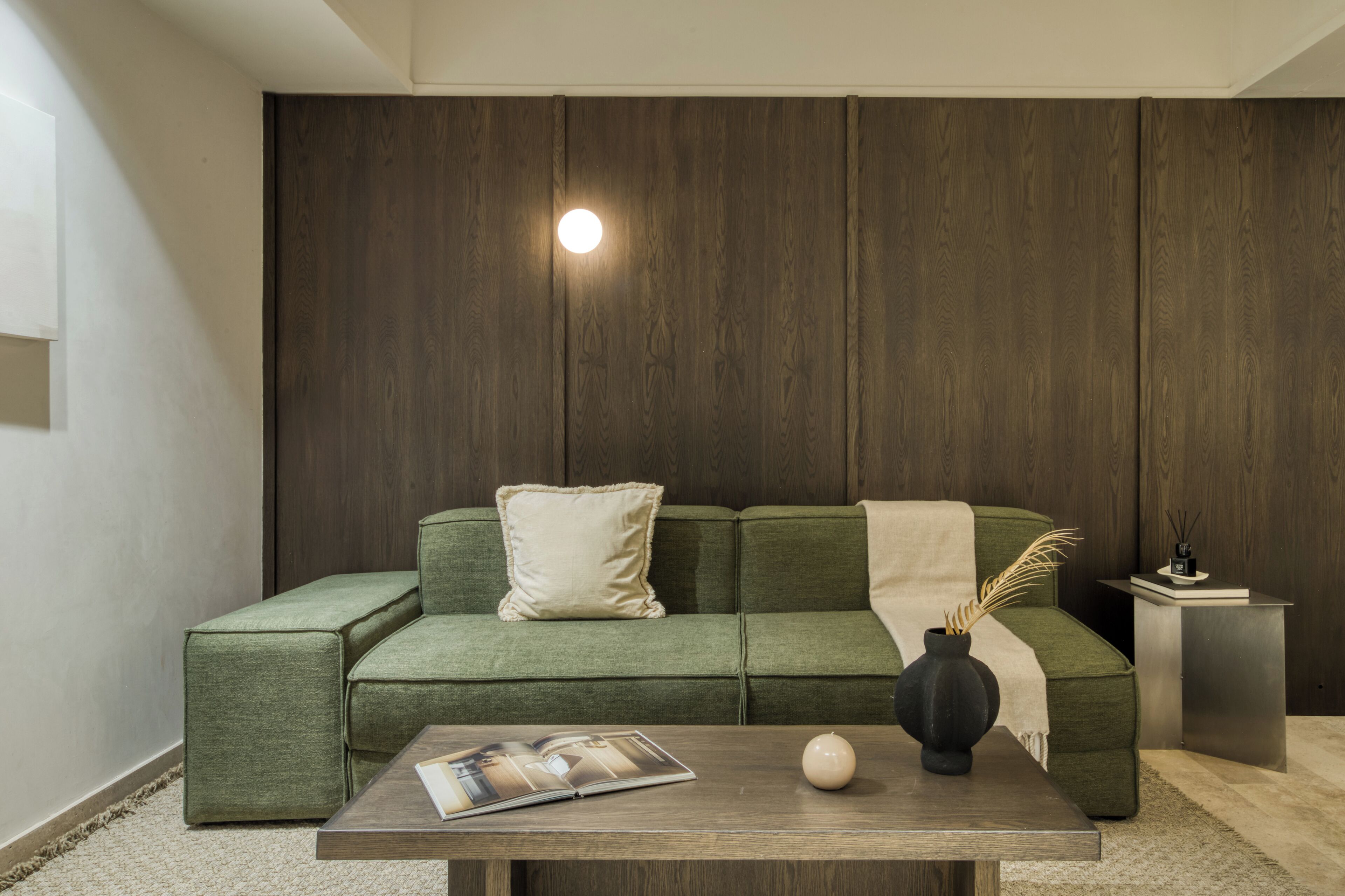 Un salon moderne avec un canapé vert, une petite table sur laquelle reposent un magazine et un vase, et un luminaire fixé au mur.