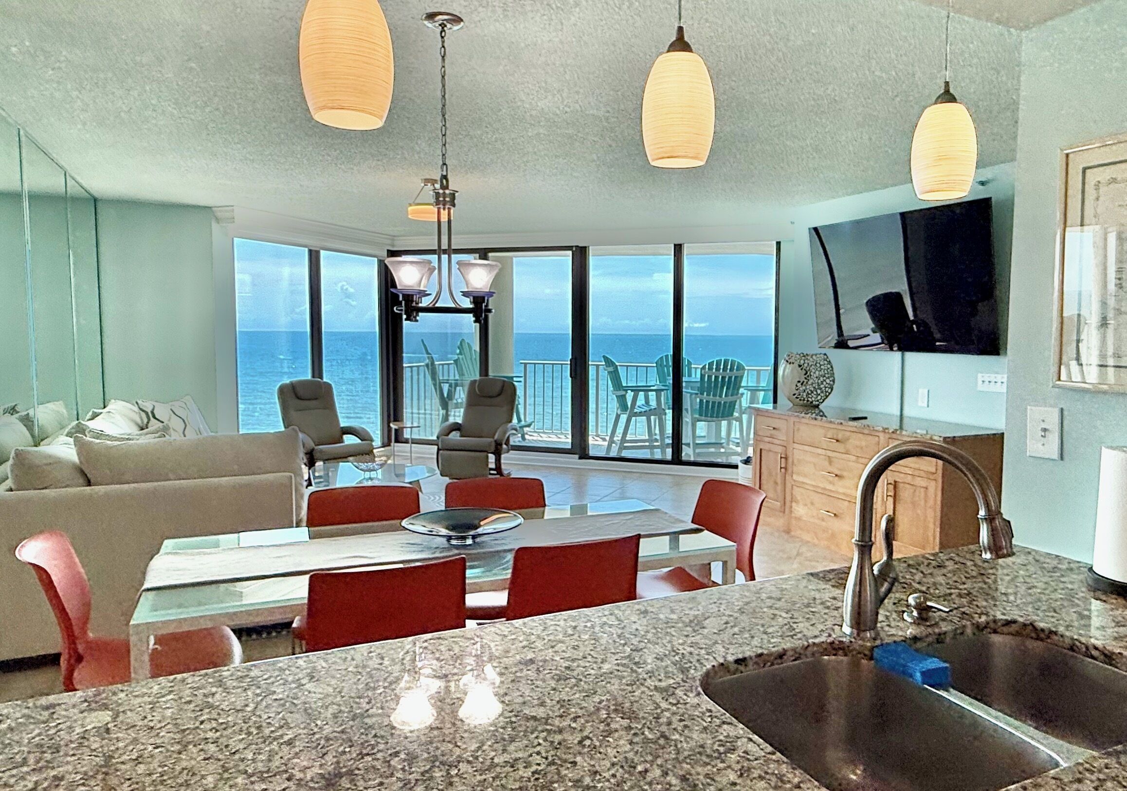 A modern kitchen with granite countertops, a stainless steel sink, and a dining area with red chairs and a glass table. There is a large window with a view of the ocean.