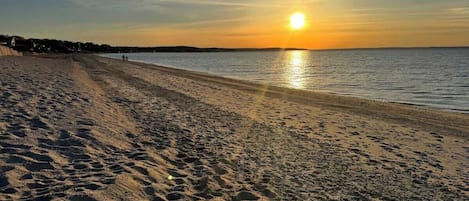 A beach with footprints leading to the water, a sunset in the background, and a clear sky.