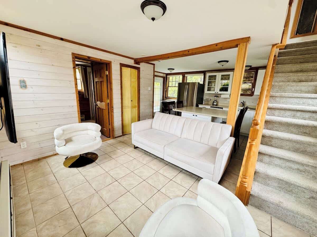 A spacious living area with a white sofa, a white armchair, and a dining table. There is a staircase leading to an upper level.
