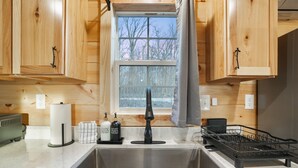 A kitchen with wooden cabinets, a stainless steel sink, a dish rack, and a window with a view of trees. - Poplar Ridge | Hocking Hills, Sleeps 7, Hot Tub (McArthur)