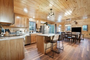 Una cocina moderna con armarios de madera, una isla central, taburetes de bar y una zona de comedor con televisión.