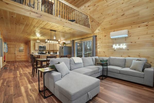 A spacious living area with a large gray sectional sofa, a dining table with chairs, a kitchen with wooden cabinets, and a balcony with a railing.