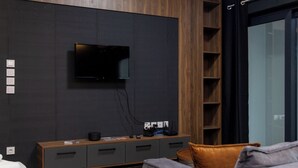 A modern living room with a flat-screen TV mounted on a dark wall, a wooden entertainment unit with drawers, and a grey sofa. - Dioroh Suites Meteora 303 (Kalabaka)