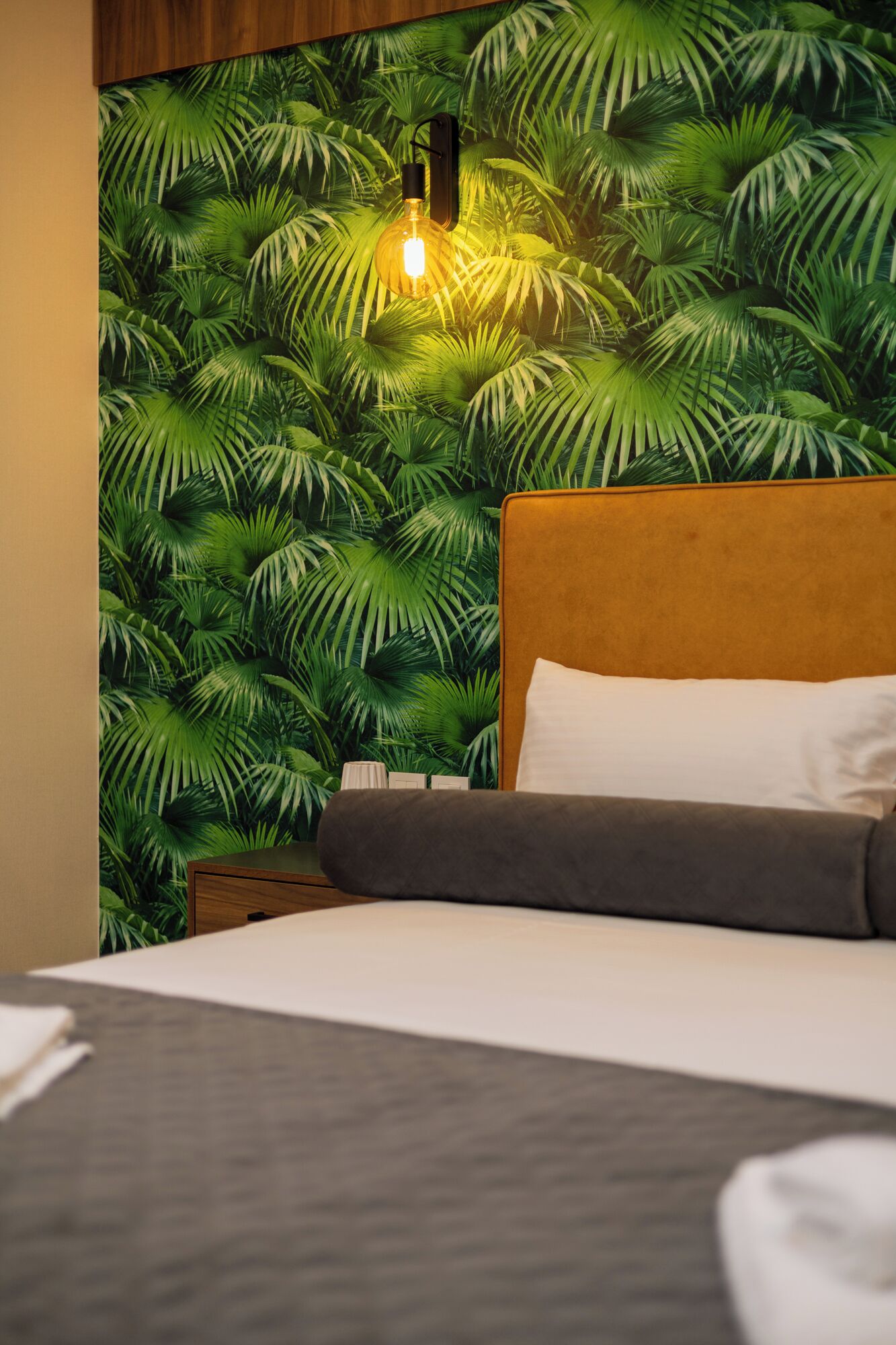 Une chambre d’hôtel avec un lit, une table de chevet et un mur orné d’un motif de feuilles tropicales.