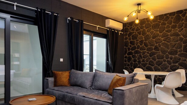 A modern living room with a grey sofa, a round wooden coffee table, and a stone wall. - Dioroh Suites Meteora 303 (Kalabaka)