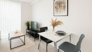 Een moderne, minimalistische woonkamer met een eethoek. Er staat een flatscreen televisie op een zwart voetstuk, een eettafel met twee stoelen en een salontafel. Op de tafel staan een potplant en een vaas met gedroogde bloemen. De kamer heeft witte muren en een groot raam met jaloezieën.