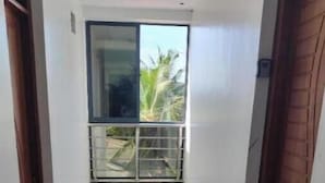 A narrow hallway with white tiled flooring, a sliding glass door leading to a balcony with a view of greenery, and wooden doors on either side.