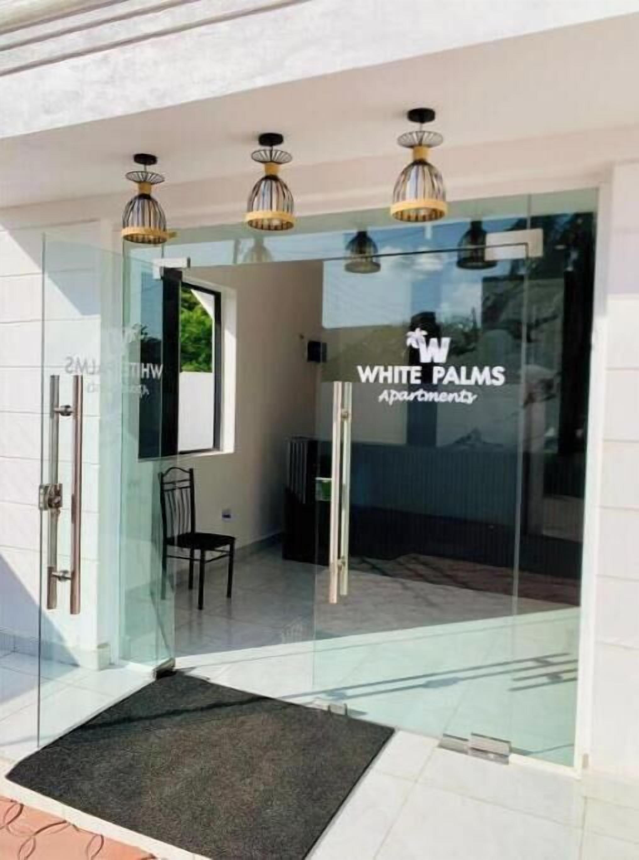 Entrada de un edificio moderno con puerta de cristal, un cartel que dice 'WHITE PALMS Apartments' y tres lámparas colgantes sobre la puerta.