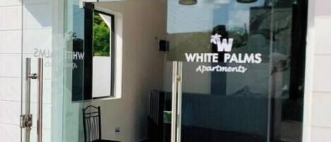 A modern building entrance with a glass door, a sign reading "WHITE PALMS Apartments," and three pendant lights above the door.