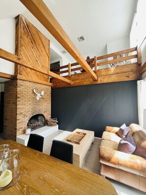 Una sala de estar acogedora con chimenea de ladrillo, vigas de madera, un sofá y una zona de comedor con mesa y sillas.