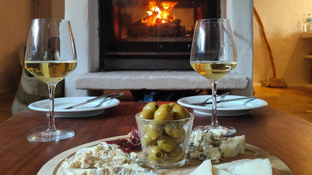 Una mesa de comedor con dos copas de vino blanco, una tabla de madera con queso, aceitunas y una rebanada de manzana, y una chimenea al fondo.