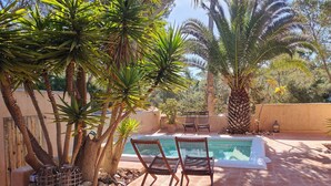 Zona de piscina con dos sillas y una mesa, rodeada de palmeras y un muro de piedra.