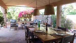 Un espacio de comedor al aire libre con techo, que incluye una mesa de madera lista para una comida, rodeada de sillas y con lámparas colgantes de tejido.