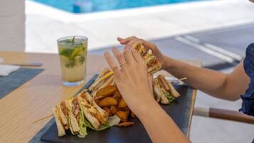 A person assembling a sandwich with a side of fries on a black plate, with a pool in the background.
