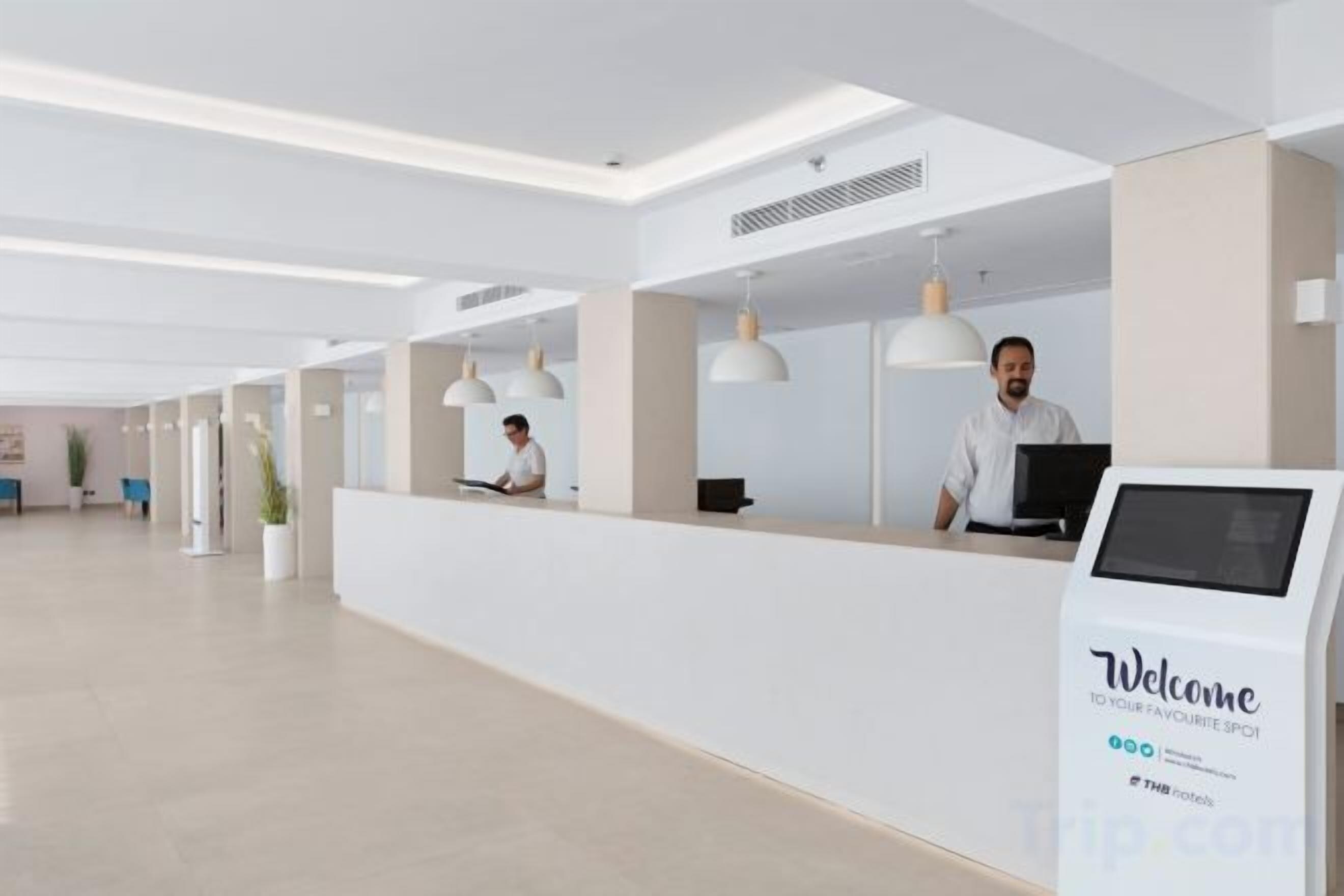 Ein Hotel-Empfangsbereich mit Empfangstresen, zwei Mitarbeitern, einem digitalen Kiosk und einer Pflanze.