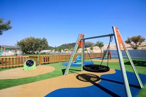 A playground with a swing set and a tire swing, surrounded by a wooden fence and a clear blue sky.