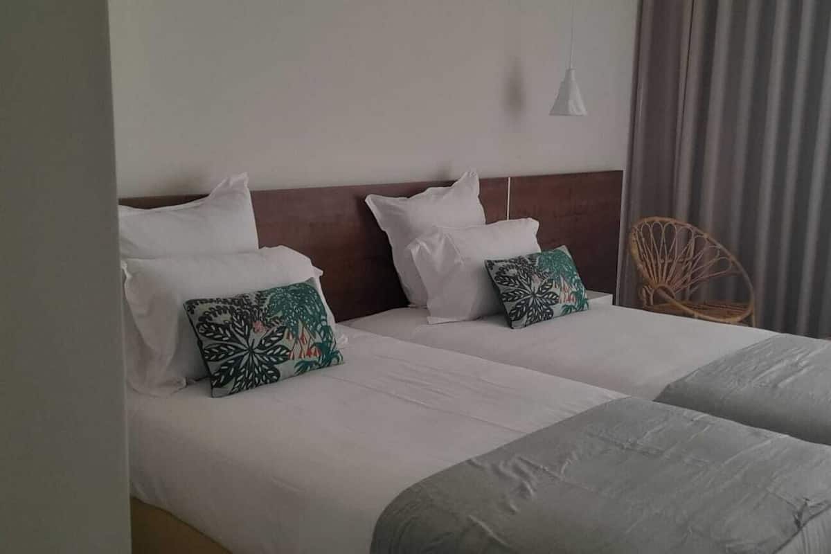 Un lit double avec du linge de lit blanc et deux coussins décoratifs, une tête de lit en bois, une chaise en osier et une fenêtre avec des rideaux.