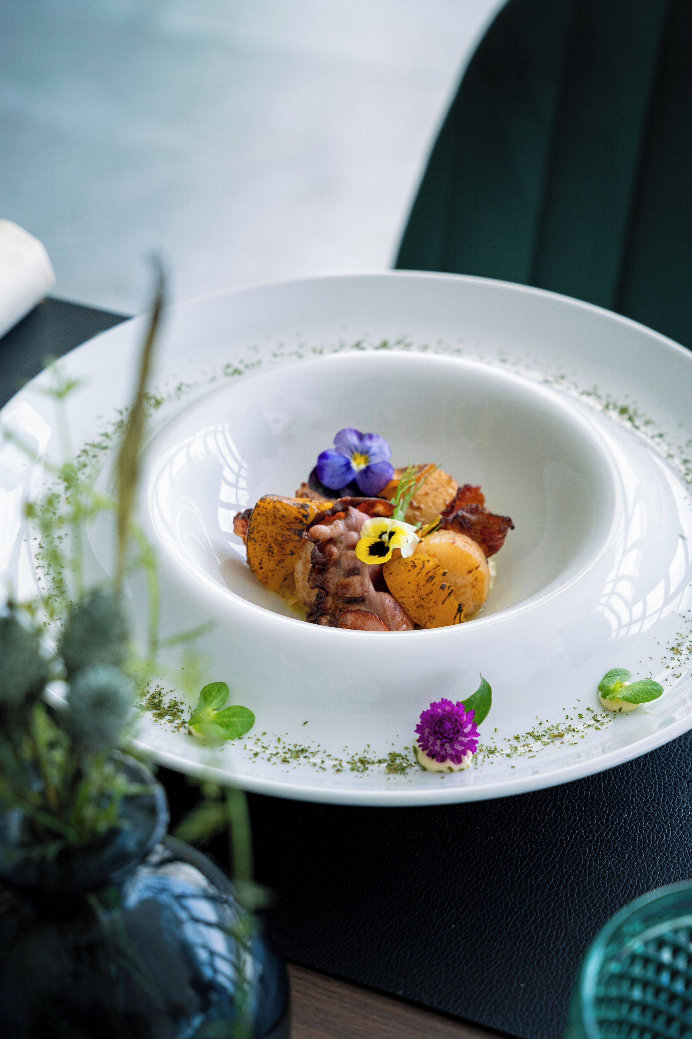 Un plato blanco con un guiso de comida, decorado con flores moradas y hojas verdes, sobre una mesa oscura.