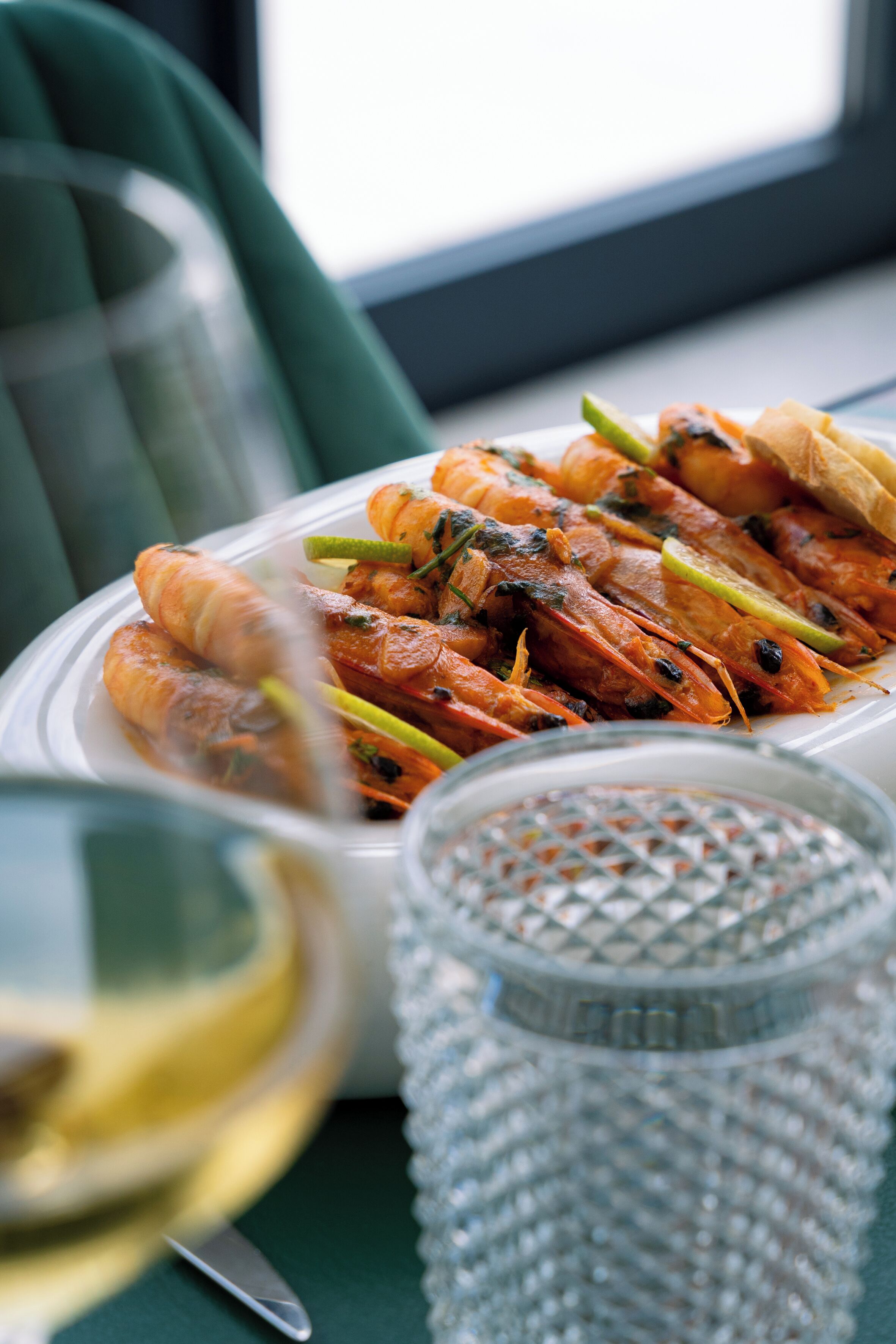 A plate of grilled shrimp with lemon slices and herbs, a glass of white wine, and a textured glass container on a table.
