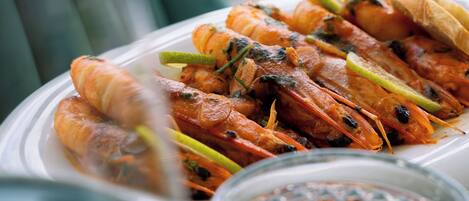 A plate of grilled shrimp with lemon slices and herbs, a glass of white wine, and a textured glass container on a table.