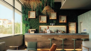 Un espace bar avec des comptoirs en bois, des plantes vertes et des tableaux encadrés. On y trouve des tabourets de bar et un coin salon avec des chaises.