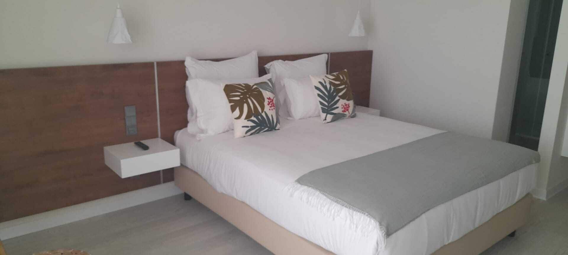 A neatly made bed with white bedding and decorative pillows, a bedside table with a remote control, and a modern headboard.